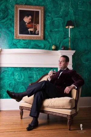 Sven Raphael Schneider sitting in burgundy velvet dinner jacket in front of green wall with a drink in hand
